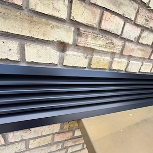 Peut inclure: Un couvercle de ventilation rectangulaire noir avec des lattes horizontales. Le couvercle est posé contre un mur de briques patinées. Le couvercle de ventilation est fini mat foncé.