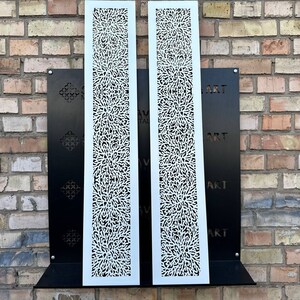 May include: Two white metal panels with a decorative cut-out design. The panels are mounted on a black metal frame against a brick wall. The text "ART" is visible on the frame.