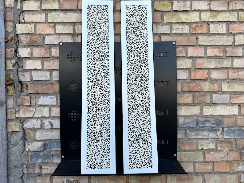 May include: Two white metal panels with a decorative cut-out design. The panels are mounted on a black metal frame against a brick wall. The text "ART" is visible on the frame.