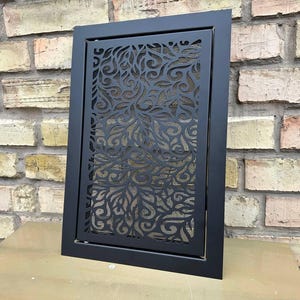 May include: Black metal vent cover with a decorative floral pattern. The rectangular cover has a solid black frame and a mesh backing. The vent is displayed against a brick wall and a wooden surface.