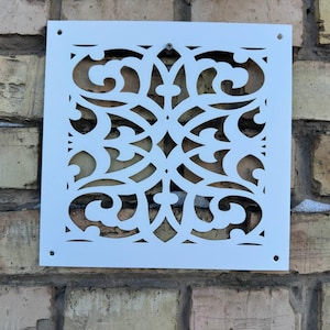 May include: A square, white decorative vent cover with an intricate, openwork design. The pattern features swirling, symmetrical shapes. The cover is mounted on a weathered brick wall, providing a contrast of textures and colours.