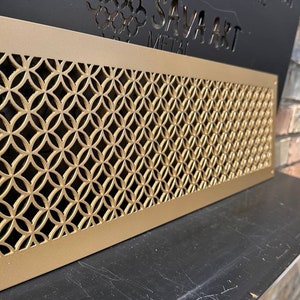 Puede incluir: Rejilla de ventilación de metal dorado con un patrón geométrico. La rejilla de ventilación es rectangular y tiene un borde elevado. El patrón está formado por círculos y óvalos superpuestos.
