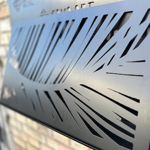 Puede incluir: Arte de pared de metal negro con un diseño recortado de una hoja de palma. El arte está montado en una pared de ladrillo.