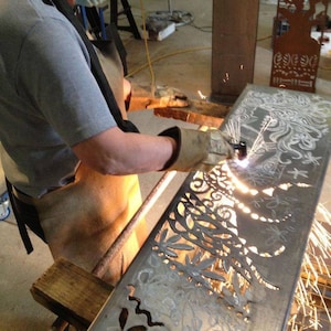 Könnte beinhalten: Ein Metallarbeiter verwendet einen Schweißbrenner, um komplizierte Blumen- und abstrakte Designs in ein rechteckiges Metallblech zu schneiden. Das Blech ist silberfarben, und Funken fliegen, während der Brenner durch das Metall schneidet. Der Arbeiter trägt Schutzkleidung.