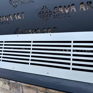 May include: A rectangular, light gray metal vent cover with horizontal slats. The vent cover is positioned in front of a black metal panel with the text "SAVA ART METAL" cut out. The vent cover has several screw holes.