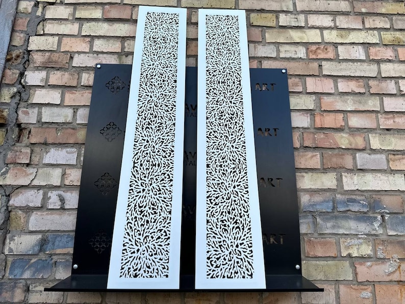 May include: Two white metal panels with a decorative, intricate, floral pattern. The panels are mounted on a black background with the word "ART" repeated three times.