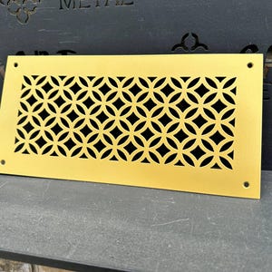 May include: A rectangular, gold-colored metal vent cover with a geometric pattern of interlocking circles and diamond shapes. The vent cover has a matte finish and is designed with four screw holes in each corner.