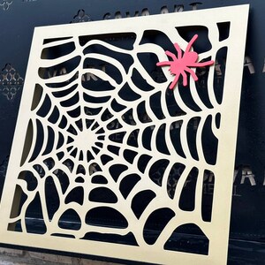 May include: A gold metal cutout of a spider web with a red plastic spider on it. The cutout is mounted on a black surface.