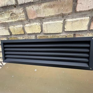May include: A rectangular, black metal vent cover with horizontal slats. The vent is mounted on a light brown surface, with a brick wall in the background. The vent has a simple, modern design.