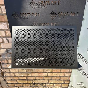 May include: A black metal decorative panel featuring a repeating geometric design. The panel is rectangular with circular and cross-shaped cutouts. The words "SAVA ART METAL" are visible in the background.