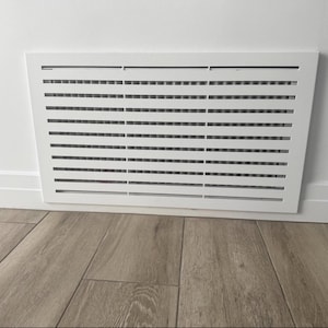 May include: A white rectangular air vent cover with horizontal slats is mounted on a white wall. The vent is positioned above a light brown wood floor. A white electrical outlet with a small white device is visible on the wall.
