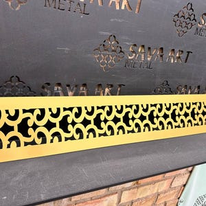 May include: A gold-colored metal decorative panel with a repeating scroll design. The panel is rectangular and has a cutout pattern. The background features the text "SAVA ART METAL" in a dark color.