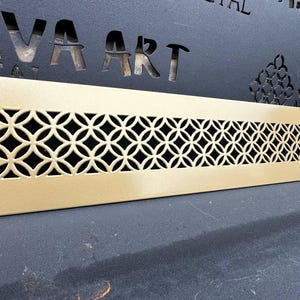 Puede incluir: Una rejilla de ventilación de metal dorado con un patrón geométrico decorativo. La rejilla de ventilación es rectangular y tiene una superficie lisa.