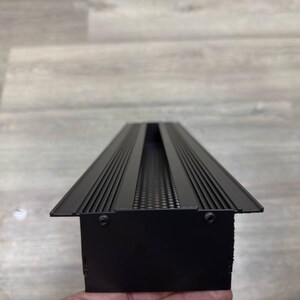 May include: Black metal light fitting with a rectangular shape and a long, narrow opening in the centre. The opening has a series of small holes along the sides.