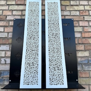 May include: Two white metal panels with a decorative, intricate, floral pattern. The panels are mounted on a black background with the word "ART" repeated vertically.