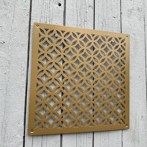 Puede incluir: Panel decorativo cuadrado de color dorado con un patrón circular repetitivo. El panel es de metal y tiene una serie de pequeños agujeros recortados para crear el diseño. Está montado sobre una superficie de madera blanca desgastada.