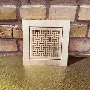 Peut inclure: Un couvercle de ventilation métallique carré avec un motif géométrique. Le couvercle est de couleur bronze clair et possède un centre en retrait à motifs. Le fond est une surface dorée texturée et un mur de briques.