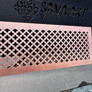 May include: A copper-colored metal vent cover with a repeating quatrefoil pattern. The rectangular cover has a border with screw holes. The words "SAVA ART METAL" are cut out of a black surface in the background.