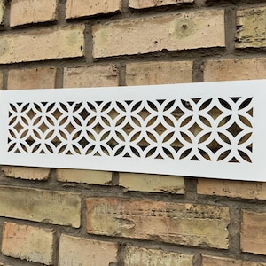May include: A white rectangular decorative vent cover with a geometric pattern of interlocking circles and diamond shapes. The vent cover is mounted on a weathered brick wall, providing a contrast in textures and colors.