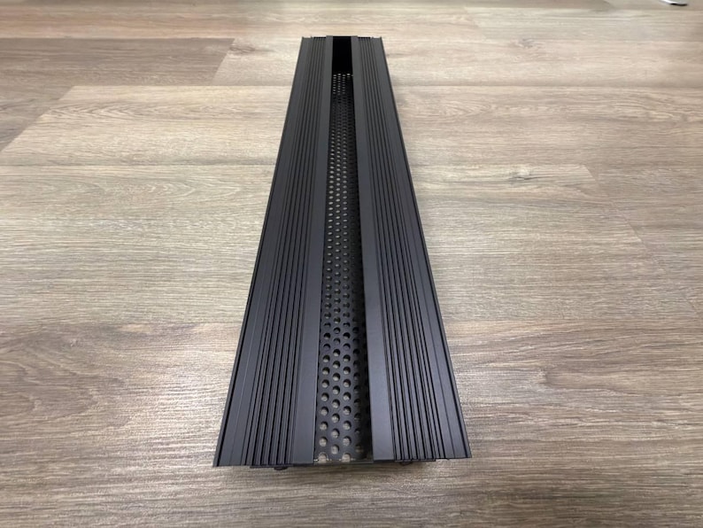 May include: Black metal vent cover with a long, narrow rectangular shape and a series of vertical lines on the surface. The vent cover has a perforated metal insert in the centre.