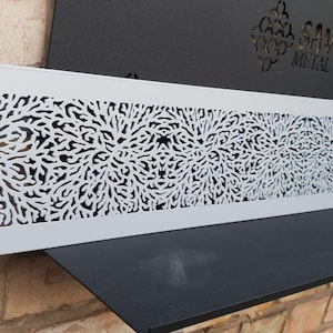 May include: A white metal vent cover with a decorative cutout design of black floral patterns. The vent cover is approximately 45 cm long.
