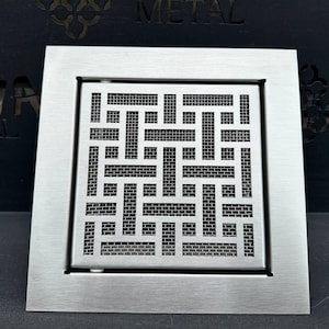 以下が含まれることがあります： 幾何学模様の正方形のつや消し金属製排水溝カバー。デザインは、相互接続された一連の長方形と正方形を特徴とし、メッシュのような外観を作り出しています。カバーは暗い背景に配置されています。