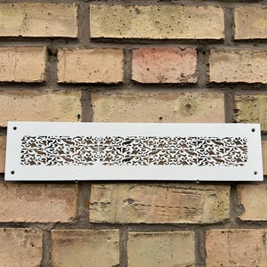 May include: A white rectangular vent cover with a decorative, swirling pattern. The vent cover is mounted on a brick wall. The pattern is cut out of the metal, allowing air to pass through.
