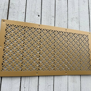 Puede incluir: Panel decorativo rectangular dorado con un patrón circular repetido. El panel tiene pequeños agujeros alrededor para la fijación. El panel metálico es probablemente para ventilación o decoración, mide aproximadamente 60 cm x 30 cm.