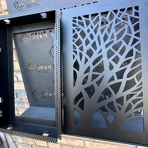 Op de afbeelding: Zwarte metalen wandkunst met een boomtakontwerp. Het kunstwerk bestaat uit twee delen: een rechthoekig frame en een paneel met een uitgesneden boompatroon. De tekst "SAVA METAL" is zichtbaar.