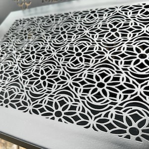 May include: A white metal vent cover with a decorative cutout design of repeating black floral patterns.