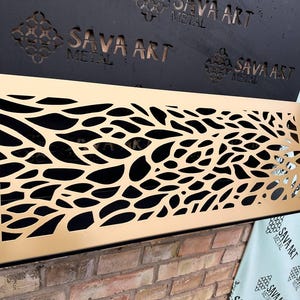 May include: A gold metal decorative vent cover with a cut-out design of overlapping leaves. The vent cover is mounted on a brick wall.