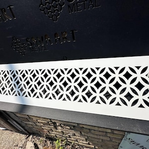 Puede incluir: Rejilla de ventilación decorativa de metal blanco con un patrón geométrico de círculos y diamantes entrelazados. La rejilla es rectangular y se asienta sobre una superficie de ladrillo. El texto "SAVA ART METAL" es visible.