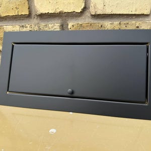 May include: A rectangular, matte black cabinet door with a recessed panel and a small, round knob. The door is set within a matching frame. The background features a weathered brick wall and a light-colored surface.