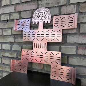 Puede incluir: Una escultura de pared de metal color cobre, plana, con forma de figura humana estilizada. La pieza presenta patrones complejos cortados con láser en todo el cuerpo, brazos y piernas, con un diseño de cabeza semicircular con motivos grabados.