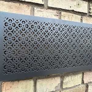 Peut inclure: Une grille de ventilation rectangulaire en métal gris foncé avec un motif décoratif répété de croix et de cercles. La grille est fixée sur un mur de briques et comporte quatre trous de vis dans les coins.