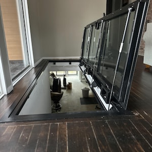 May include: A black metal door with a glass panel opens to reveal a basement space. The door is hinged at the top and opens upwards. The basement space is visible through the glass panel.