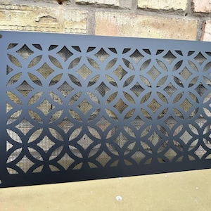 Puede incluir: Panel decorativo de metal negro con un patrón geométrico de círculos y rombos entrelazados. El panel tiene un respaldo de malla y es rectangular con esquinas redondeadas. Tiene cuatro orificios de montaje.