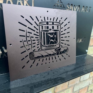 Puede incluir: Una pieza de arte de pared de metal con un diseño recortado de un vaso de whisky con hielo y un cigarro. El diseño está rodeado por un patrón de rayos de sol.