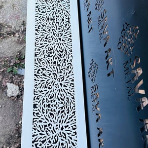 May include: A white metal panel with a decorative cut-out design. The design features a repeating pattern of small, intricate shapes that resemble branches or leaves. The panel is mounted on a brick wall next to a black metal panel with the text "SAVA ART METAL" repeated multiple times.