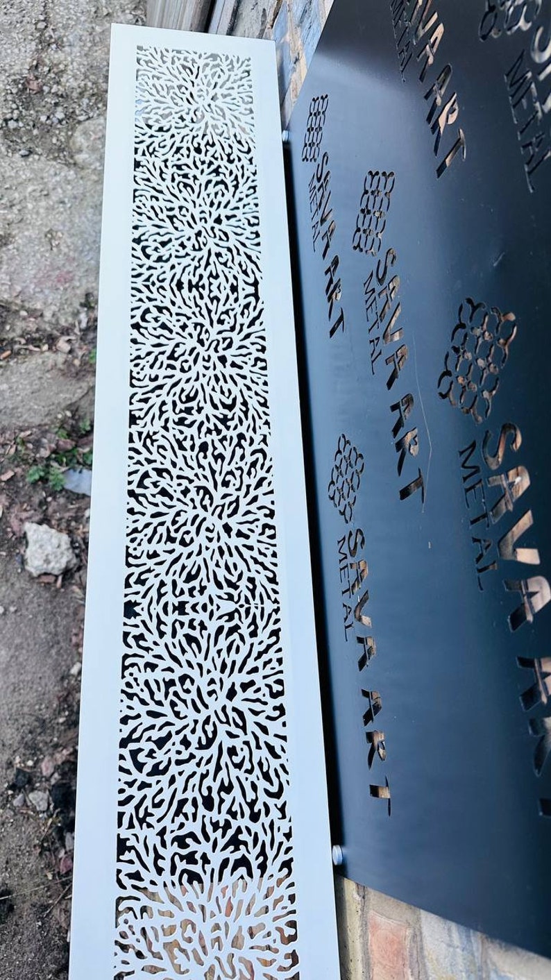 May include: A white metal panel with a decorative cut-out design. The design features a repeating pattern of small, intricate shapes that resemble branches or leaves. The panel is mounted on a brick wall next to a black metal panel with the text "SAVA ART METAL" repeated multiple times.