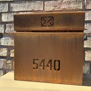 May include: A rectangular, weathered metal mailbox with the number "5440" cut out on the front. The top section features a cut-out envelope symbol. The mailbox has a rustic, aged appearance, set against a brick wall.