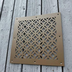 May include: A square, gold-colored metal vent cover featuring a repeating geometric pattern of interlocking circles. The flat panel has a solid border with pre-drilled screw holes positioned along the top, bottom, and sides for mounting purposes.