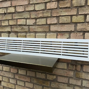 May include: A white rectangular vent cover with horizontal slits is mounted on a brick wall. Below it is a dark, rectangular shelf with a gold-colored trim. The brick wall has a mix of yellow, brown, and red bricks.