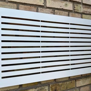 Puede incluir: Una cubierta de ventilación rectangular blanca con ranuras negras horizontales. La ventilación está montada en una pared de ladrillo. La cubierta de ventilación es un elemento de diseño moderno, probablemente para sistemas HVAC o similares.