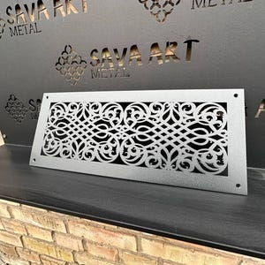 Puede incluir: Una rejilla de ventilación de metal plateado con un diseño floral decorativo e intrincado. La rejilla de ventilación es rectangular y tiene un borde ligeramente elevado. La rejilla de ventilación está hecha por Sava Art Metal.