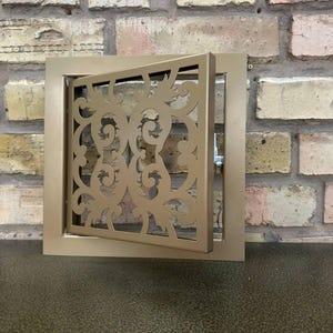 Peut inclure: Une grille de ventilation carrée en métal doré avec un motif ornemental découpé au laser. Le panneau central est monté sur charnières et légèrement ouvert, révélant un petit mécanisme de verrouillage métallique sur le côté du cadre.