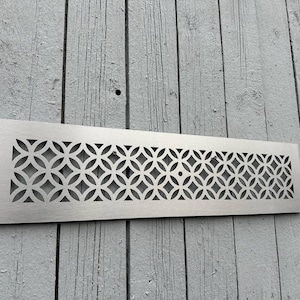 May include: A rectangular, brushed metal vent cover with a repeating geometric pattern of interlocking circles and diamond shapes. The vent cover is mounted on a weathered, gray wooden surface.