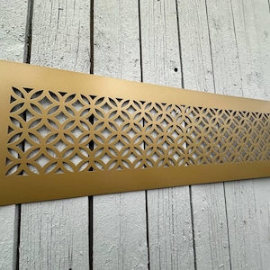 Puede incluir: Una rejilla de ventilación rectangular de metal dorado con un patrón geométrico repetitivo de círculos entrelazados. La rejilla está montada sobre una superficie de madera envejecida pintada de blanco.