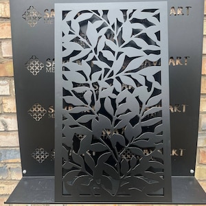 Puede incluir: Pantalla de metal negro con un diseño recortado de hojas y ramas. La pantalla está montada en un estante negro.