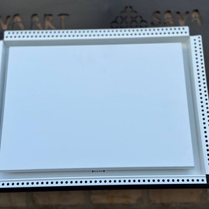 May include: White drywall panel with a white frame. The frame has a series of small holes around the perimeter. The panel is sitting on a black surface.